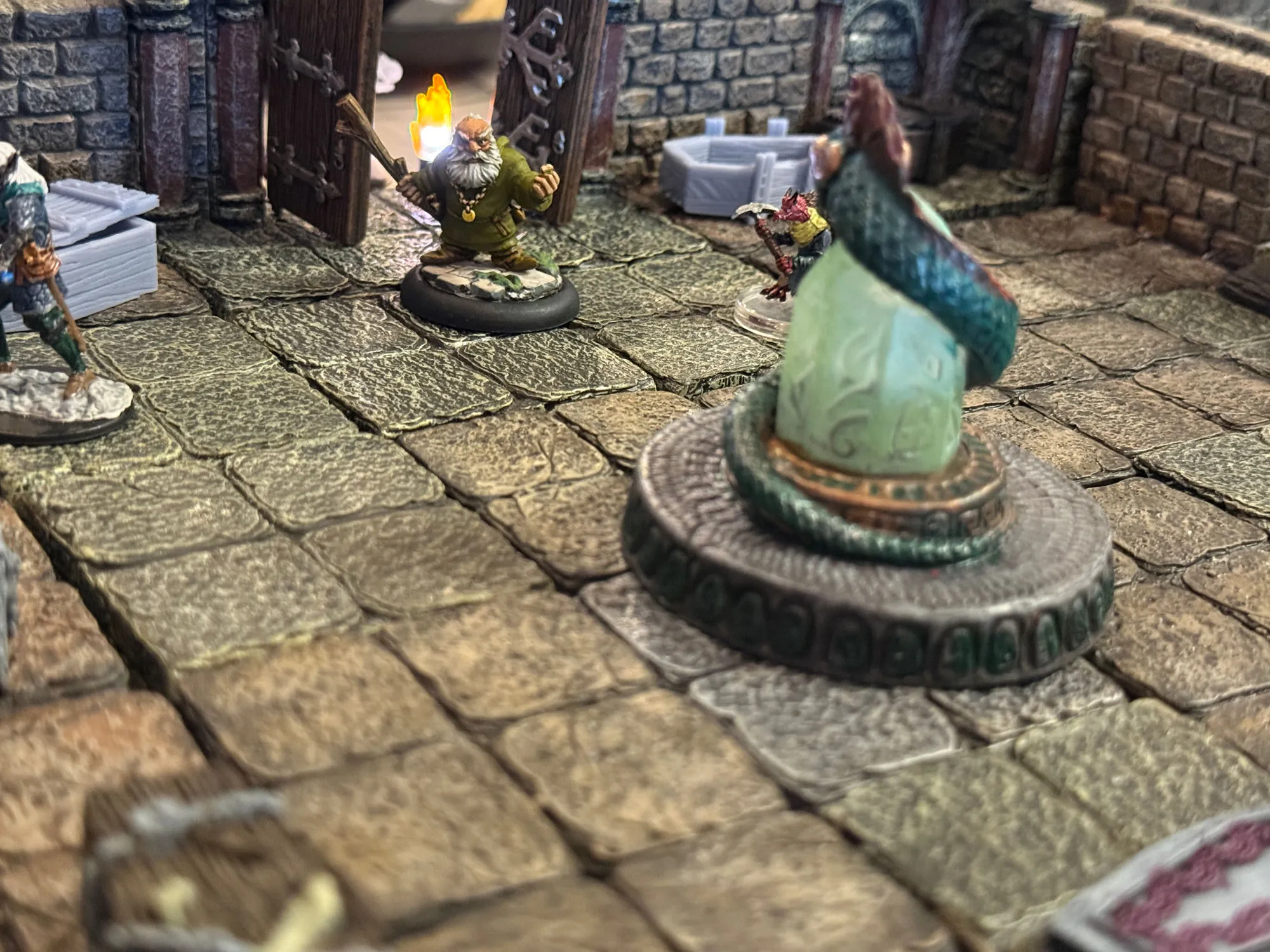 Borb, a bearded dwarf miniature in a green robe, holds up a lit torch on a flagstone dungeon floor, facing a large green crystal set on a stone dais, with another adventurer waiting in the shadows beyond it.