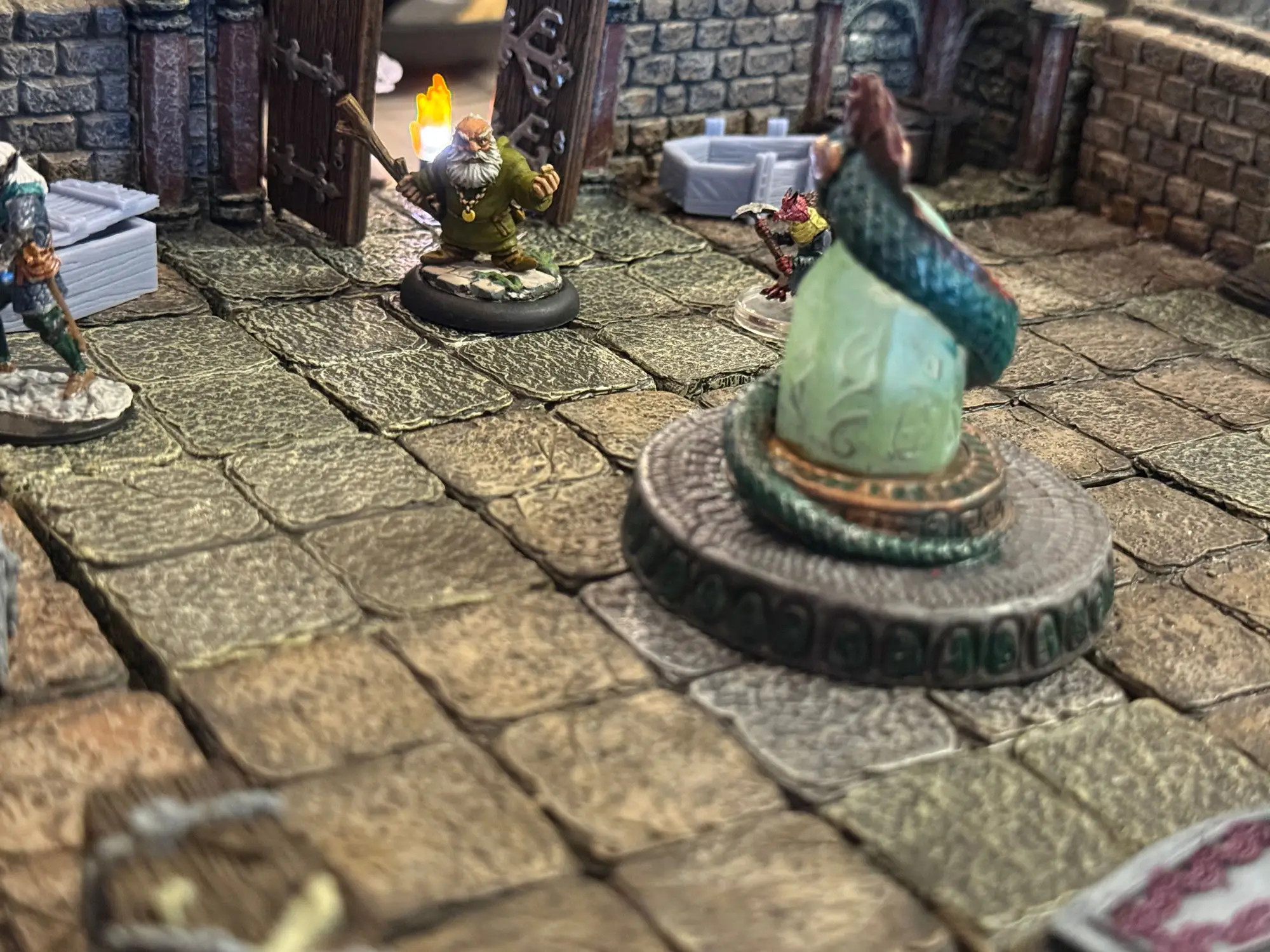 Borb, a bearded dwarf miniature in a green robe, holds up a lit torch on a flagstone dungeon floor, facing a large green crystal set on a stone dais, with another adventurer waiting in the shadows beyond it.